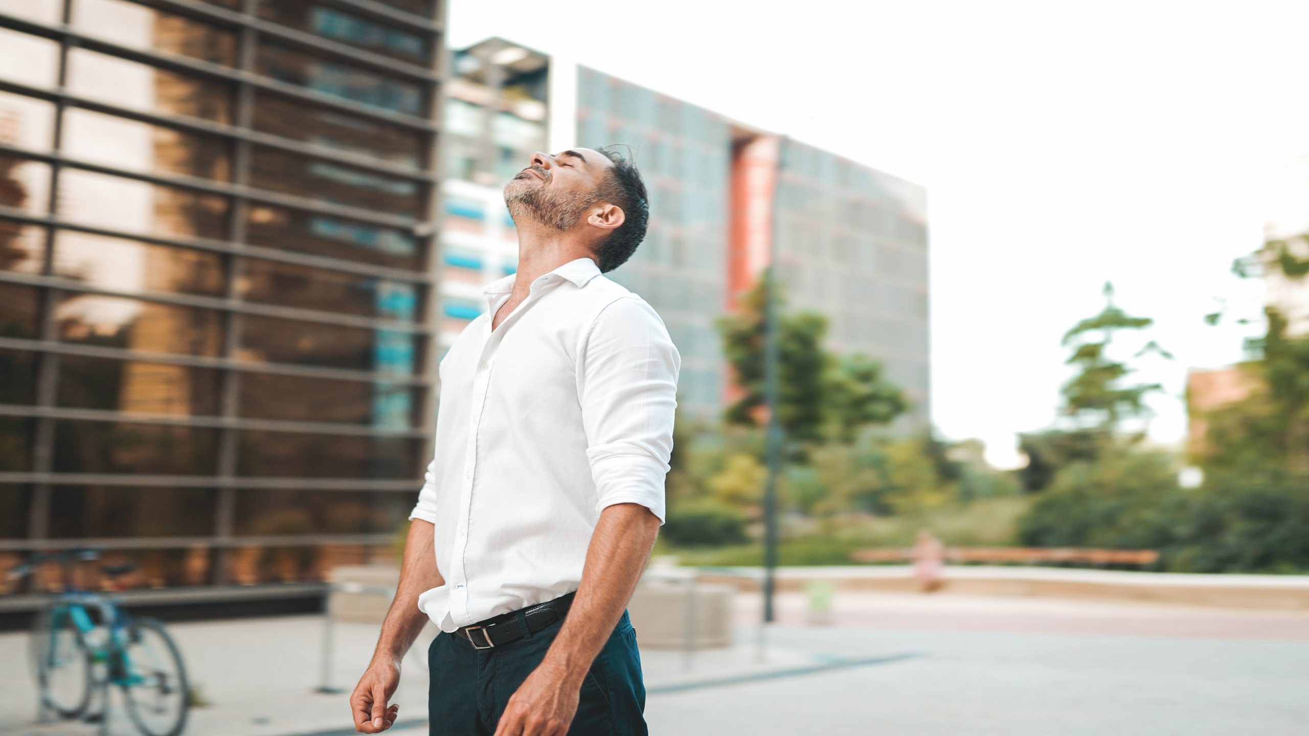 Mature businessman with neat beard, unbuttoning white shirt stands on the street of modern city. Successful man is relaxed, enjoys life.