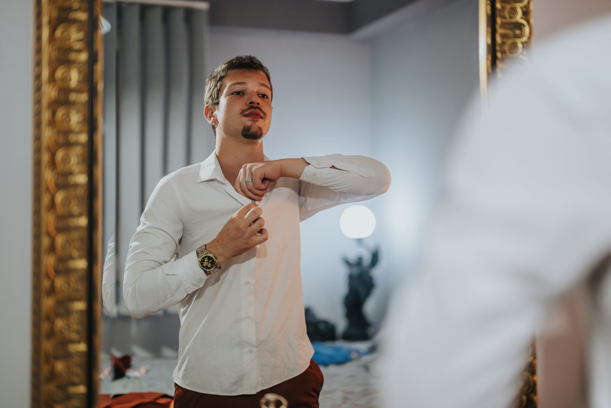 A man in a white shirt adjusts his collar while looking in the mirror, getting ready for a night out. The scene shows anticipation and attention to detail.